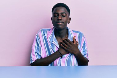 Gündelik giysiler giymiş genç Afro-Amerikan bir adam masaya oturmuş elleri göğsünde, gözleri kapalı ve yüzünde minnet dolu bir jestle gülümsüyor. sağlık kavramı. 
