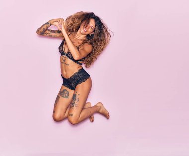 Dövmeli, iç çamaşırlı, mutlu gülümseyen genç bir kadın. Yüzünde tebessümle zıplıyor, kalp şeklinde, elleri izole pembe arka planda.