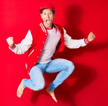 Beyzbol kıyafeti giyen genç yakışıklı bir adam. Açık ağızla zıplıyor, kırmızı arka planda yumruklar havada, kazanan işareti yapıyor.