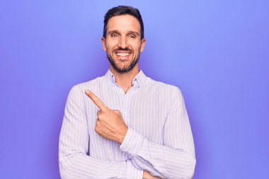 İzole edilmiş mor arka planda dikilen, sıradan çizgili tişört giyen genç, yakışıklı bir adam. Gülümseyen, eli ve parmağı yana doğru olan.