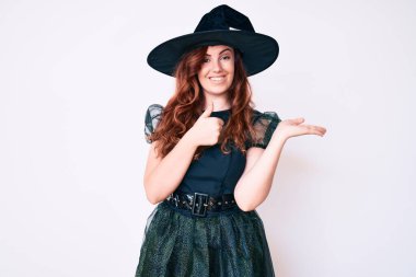 Cadı cadılar bayramı kostümü giymiş genç güzel bir kadın avuç içini gösteriyor ve baş parmaklarıyla iyi bir jest yapıyor, mutlu ve neşeli gülümsüyor. 