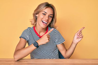 Masaya oturmuş gülümseyen ve kameraya bakan beyaz elbiseli güzel bir kadın iki eliyle ve parmaklarıyla işaret ediyor.. 