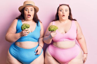 Bikini giyen genç ve büyük beden ikizler brokoli tutuyorlar. Korkutulmuş ve ağzı açık şaşırmış bir şekilde, inançsız bir yüz. 