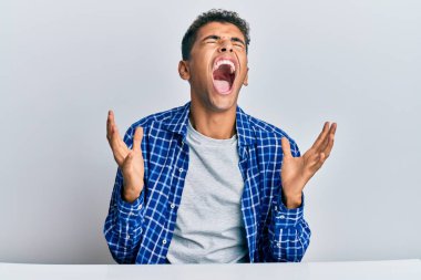 Genç ve yakışıklı Afro-Amerikan adam, gündelik giysiler içinde masaya oturmuş deli gibi bağırıyor ve agresif bir ifadeyle bağırıyor ve kollarını kaldırıyor. hüsran kavramı. 
