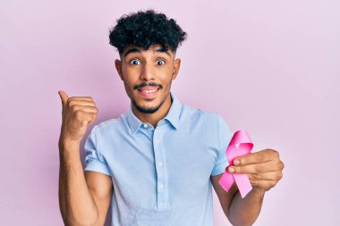 Genç Arap yakışıklı adam pembe kanser kurdelesi tutuyor başparmağıyla yan tarafa bakıyor ağzı açık gülümsüyor. 