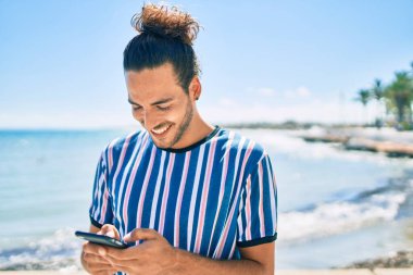 Genç İspanyol adam sahilde akıllı telefon kullanarak mutlu bir şekilde gülümsüyor..