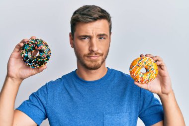 Yakışıklı beyaz adam elinde lezzetli renkli çörekler tutuyor şüpheci ve gergin, sorun yüzünden surat asıyor. negatif kişi. 