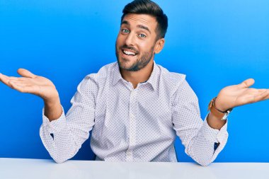 Masada oturmuş gülümseyen, iki elini de açık gösteren, sunan, reklam yapan, iş kıyafetleri giyen yakışıklı İspanyol adam. 