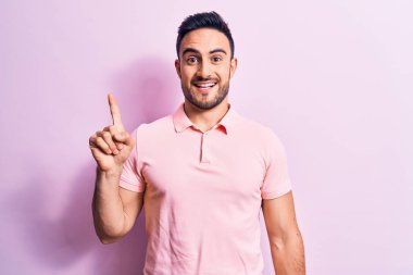 Genç, sakallı, sıradan polo giyen, pembe arka planda duran, bir fikir veya soruyla gülümseyen, mutlu bir yüzle işaret eden yakışıklı bir adam.