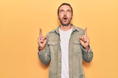 Gündelik giysiler giyen genç yakışıklı adam şaşırmış ve şaşırmış bir şekilde yukarı bakıyor ve parmaklarıyla işaret ediyor ve kollarını kaldırıyor.. 