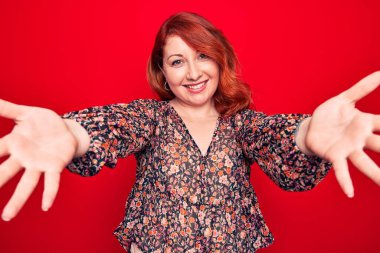 Genç, güzel, kızıl saçlı, çiçekli bir elbise giyen, kırmızı arka planda dikilmiş kameraya bakıp sarılmak için kollarını açmış gülümseyen bir kadın. Mutluluğu kucaklayan neşeli bir ifade..