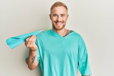 Elinde kağıt uçak tutan genç beyaz bir adam. Pozitif ve mutlu görünüyor. Kendine güvenen bir gülümsemeyle, dişlerini gösteriyor. 