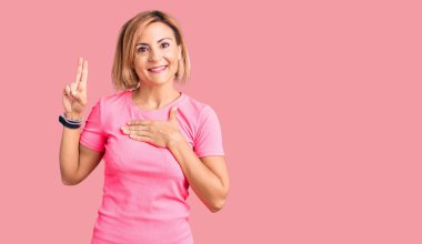 Genç sarışın kadın spor kıyafetleri giyiyor, gülümsüyor, elleri göğsünde ve parmakları havada küfür ediyor, sadakat yemini ediyor. 