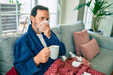 Orta yaşlı bir adam evde üşütmüş ve ateşi var. Kanepede oturan grip hastası.