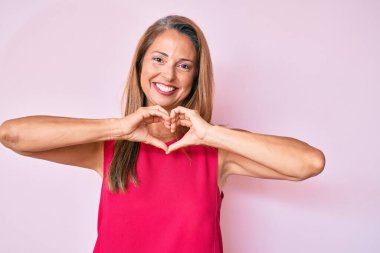 Orta yaşlı İspanyol kadın, gündelik kıyafetler giyip, aşık gülümsüyor, elleriyle kalp sembolü yapıyor. romantik kavram. 