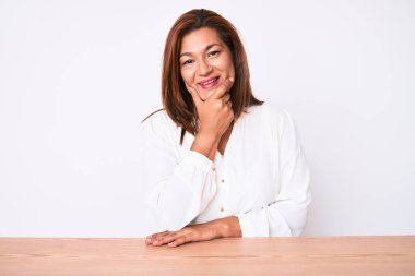 Orta yaşlı esmer, Latin iş kadını, gündelik beyaz gömleğini giymiş masaya oturmuş kameraya kendinden emin bir şekilde bakıyor kollarını kavuşturmuş ve çenesini kaldırmış gülümsüyor. Olumlu düşünmek. 