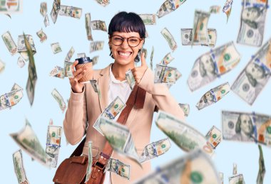 Kısa saçlı, iş çantası takan, kahvesini yudumlayan, mutlu ve pozitif gülümseyen, başparmağı mükemmel ve onay işareti olan esmer bir kadın.