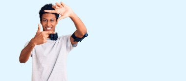 Genç Afrikalı Amerikalı adam spor kıyafetleri giyiyor ve kulaklık takıyor gülümsüyor. Elleriyle ve parmaklarıyla mutlu bir yüz çiziyor. yaratıcılık ve fotoğrafçılık kavramı. 