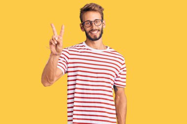 Sakallı, sıradan giyinmiş, gözlüklü, gülümseyen, parmaklarını zafer işareti yaparken gösteren sarışın yakışıklı bir adam. İki numara.. 