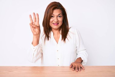 Orta yaşlı esmer, İspanyol iş kadını, gündelik beyaz gömlekli, masada oturuyor ve üç numarayı işaret ediyor, kendinden emin ve mutlu bir şekilde gülümsüyor.. 