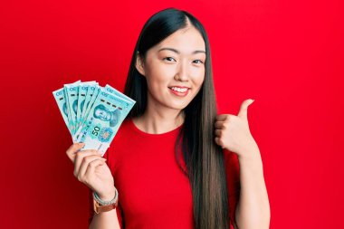 50 yuan Çin banknotları tutan genç Çinli kadın mutlu ve pozitif gülümsüyor, başparmağıyla mükemmel ve onay işareti yapıyor. 