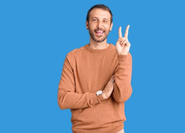 Sıradan kıyafetler giyen, gülen, kameraya göz kırpan, zafer işareti yapan genç yakışıklı bir adam. İki numara.. 