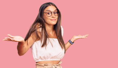 Gündelik kıyafetler giyen, gözlüklü, kafası karışmış, elleri ve kolları havada genç İspanyol bir kadın. şüphe kavramı. 