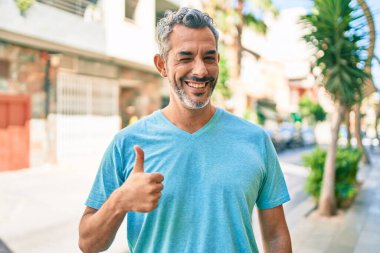 Şehrin caddesinde günlük giysiler giymiş, mutlu ve pozitif gülümseyen orta yaşlı, gri saçlı bir adam. Başparmağıyla mükemmel ve onay işareti yapıyor. 