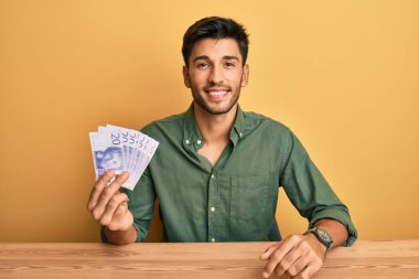 Elinde 20 İsveç banknotu tutan yakışıklı genç adam pozitif ve mutlu görünüyor ve kendine güvenen bir gülümsemeyle dişlerini gösteriyor. 