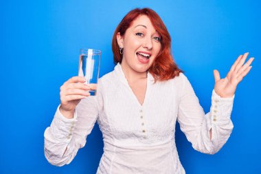 Genç ve güzel kızıl saçlı kadın mavi arka planda bir bardak su içerek başarıyı mutlu bir gülümsemeyle ve kazanan ifadeyle el kaldırarak kutluyor.