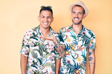 Genç eşcinsel iki adam yaz şapkası ve Hawaii gömleği giyip neşeli bir şekilde gülümsüyor ve avuç içiyle kameraya bakıyor.. 