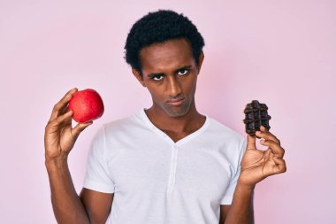 Kırmızı elmalı ve çikolatalı waffle tutan yakışıklı Afrikalı adam şüpheci ve gergin, sorun yüzünden surat asıyor. negatif kişi. 