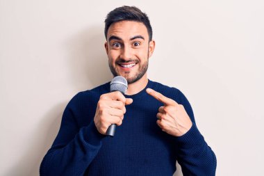 Sakallı genç yakışıklı şarkıcı beyaz arka planda mikrofon kullanarak şarkı söylüyor.