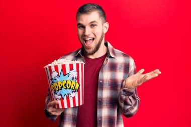 Patlamış mısır yiyen genç yakışıklı adam başarıyı mutlu bir gülümsemeyle kutluyor ve kazanan yüz ifadesi kaldırıyor. 