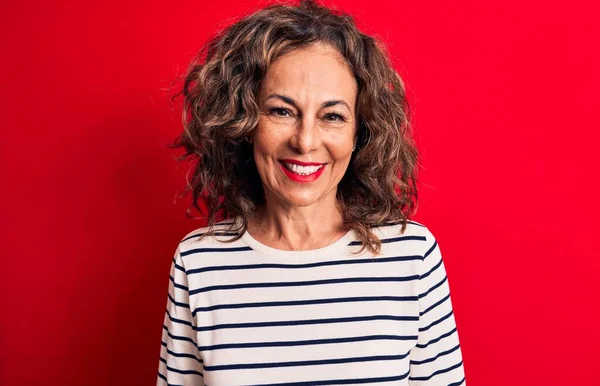 Orta yaşlı, esmer, çizgili tişört giyen güzel bir kadın kırmızı arka planda mutlu ve havalı bir gülümsemeyle dikiliyor. Şanslı kişi..