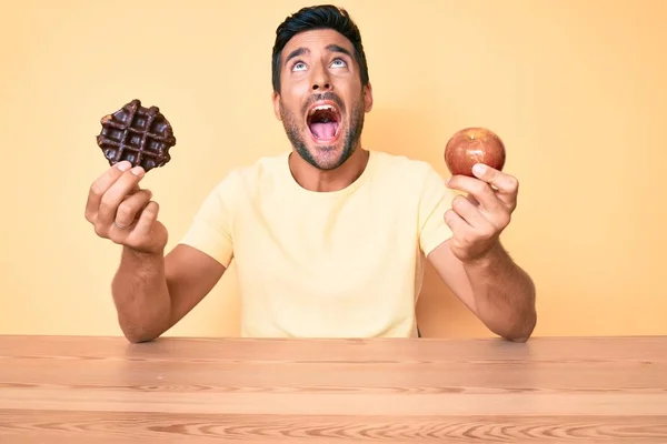 チョコレートのワッフルを保持して朝食を食べる若いヒスパニック系の男とアップル怒りと怒りの叫び 怒りで叫んで検索アップ — ストック写真
