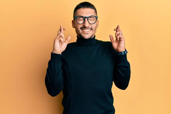 Handsome man with tattoos wearing turtleneck sweater and glasses gesturing finger crossed smiling with hope and eyes closed. luck and superstitious concept.
