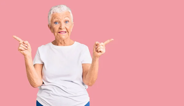 年长的漂亮女人 蓝眼睛 灰头发 穿着随意的白色T恤 自信地微笑着 用手指指向不同的方向 复制广告空间 — 图库照片