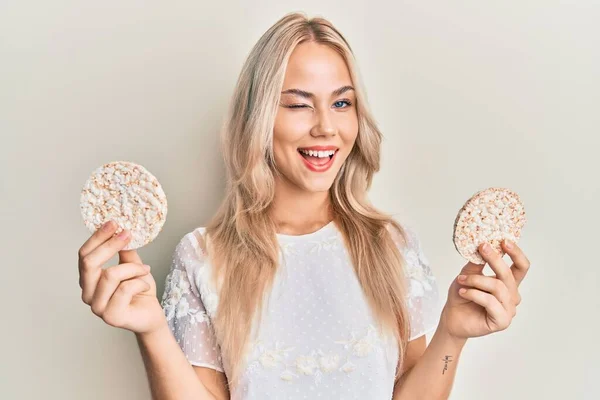 Beautiful caucasian blonde girl eating healthy rice crackers winking looking at the camera with sexy expression, cheerful and happy face. — Stock Image Beautiful Caucasian Blonde Girl Eating Healthy Rice Crackers Winking Looking Royalty Free Stock Photos