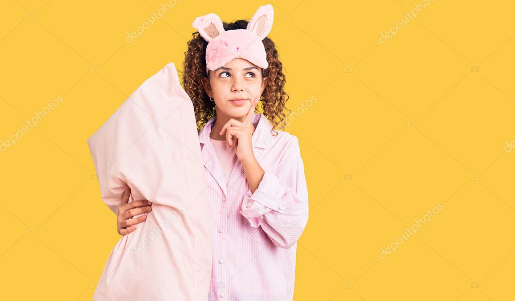 Beautiful kid girl with curly hair wearing sleep mask and pajamas holding pillow serious face thinking about question with hand on chin, thoughtful about confusing idea 