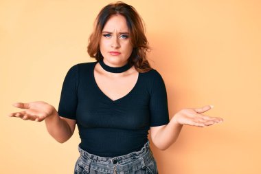 Gündelik kıyafetler giyen, kafası karışık, kolları ve elleri havada genç, beyaz bir kadın. şüphe kavramı. 