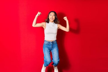 Genç, güzel Çinli kadın rahat kıyafetler giyip mutlu gülümsüyor. Yüzünde tebessümle zıplıyor, kırmızı arka planda yumruklarıyla kazanan jestler yapıyor.