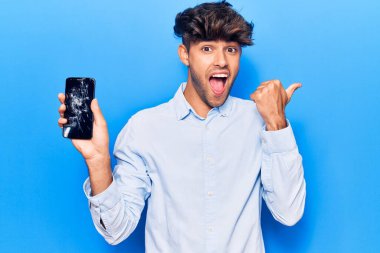 Kırık akıllı telefon tutan genç İspanyol adam kırık ekranı gösteriyor. Parmağını yan tarafa uzatıp gülümsüyor ve ağzı açık. 