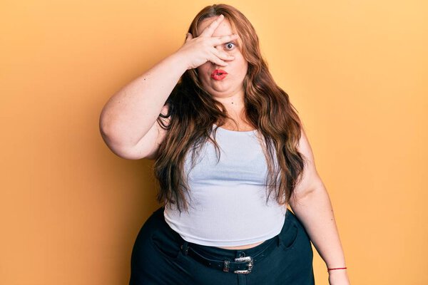 Plus size caucasian young woman wearing casual clothes peeking in shock covering face and eyes with hand, looking through fingers afraid 