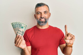 Pohledný muž středního věku drží 20 dolarů bankovky s úsměvem s nápadem nebo otázkou ukazující prstem se šťastnou tváří, číslo jedna 