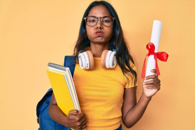 Sırt çantası takan, diplomalı, komik yüzlü, genç Hintli bir kız. Ağzı havayla şişer, havayı yakalar.. 