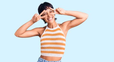 Kısa saçlı, kısa saçlı, barış sembolü yapan, yüzünde gülümseme ile zafer gösteren genç esmer kadın. 