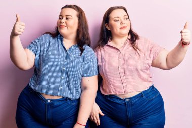 Genç ve cüsseli ikizler gündelik kıyafetler giyip gururlu görünüyor, gülümsüyor ve yan tarafa baş parmak hareketi yapıyorlar. 