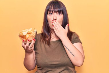 Genç ve cüsseli bir kadın elinde patates cipsi tutarak ağzını eliyle kapatıyor, şok geçiriyor ve hata yapmaktan korkuyor. şaşırmış ifade 