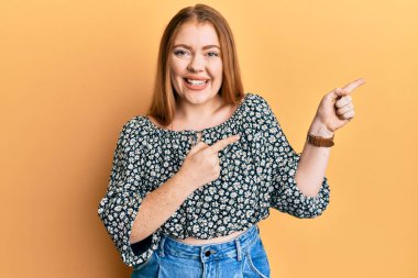 Çiçek desenli gömlek giyen güzel, kızıl saçlı bir kadın gülümseyip kameraya bakıyor ve iki eliyle ve parmaklarıyla kenarı işaret ediyor.. 